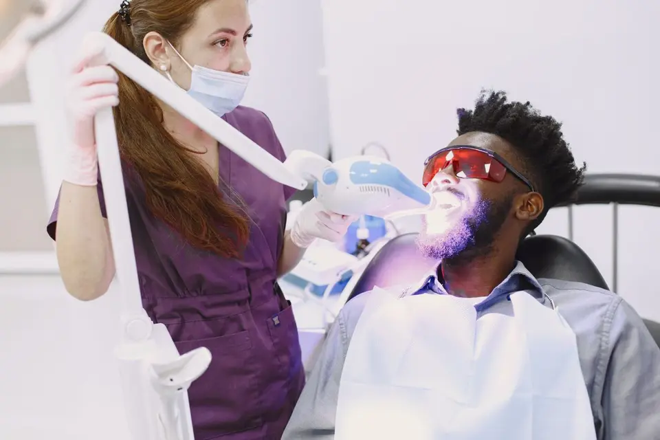 Dentist performing teeth whitening procedure on patient with specialized equipment.