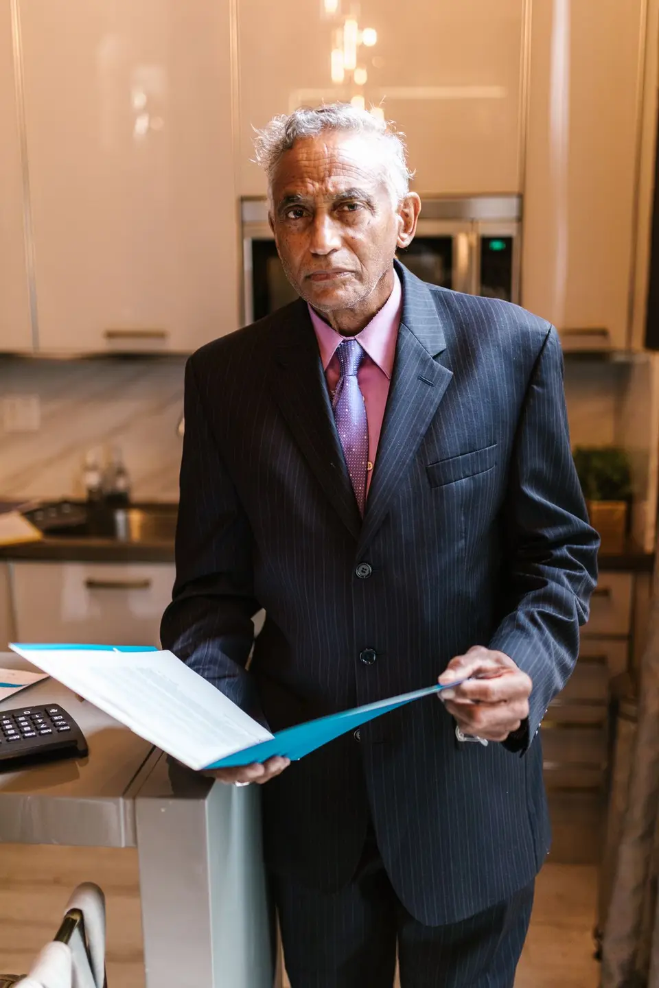 Elderly businessman in a suit holding documents in a modern kitchen setting.