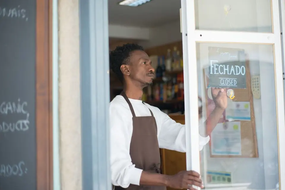 African man closing a store with a sign in Portuguese, wearing an apron.